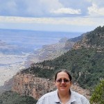 Smiling woman with brown hair and sunglasses standing by the Grand Canyon.