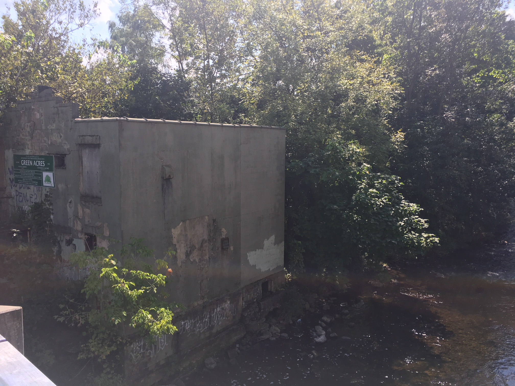 An old crumbling building sits on the shore of a river that is flowing to the right. Trees and shrubs are growing around the foundation.