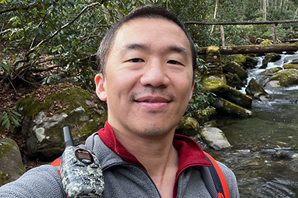ShiMing Huang stands with a backdrop of a rushing stream and rustic trail bridge.