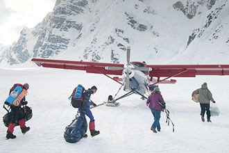 A group carrying packs walks toward an airplane on a glacier with snowy mountains in the background.