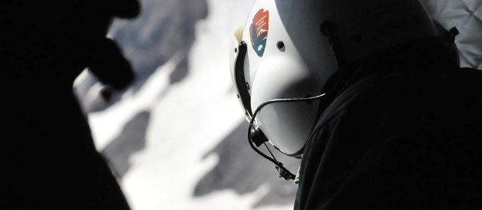 person in a flight helmet looks out at a snowy mountainous landscape from a helicopter.