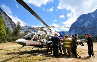 A group loads a stretcher into a helicopter in a meadow surrounded by mountains.