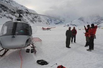 group stands outside of helicopter on snowy field with an airplane in the background