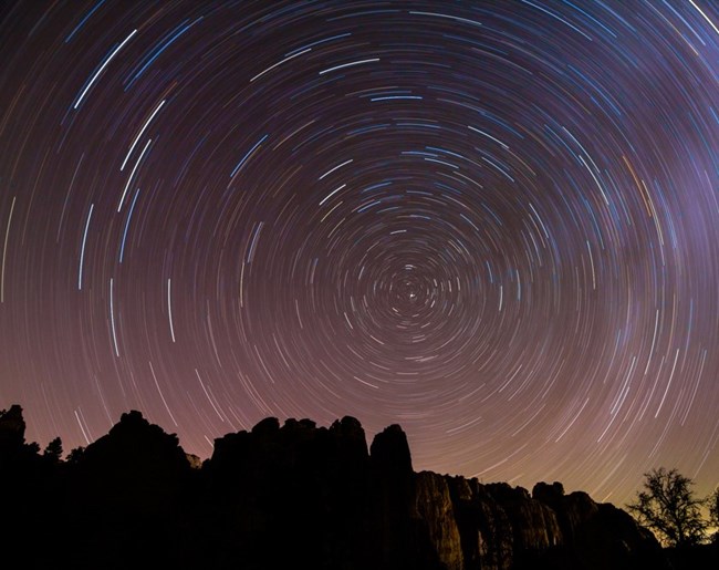 A long exposure photo of the night sky showing star trails centered around one star. The sky fades from blue to orange, and mountains are in the foreground.