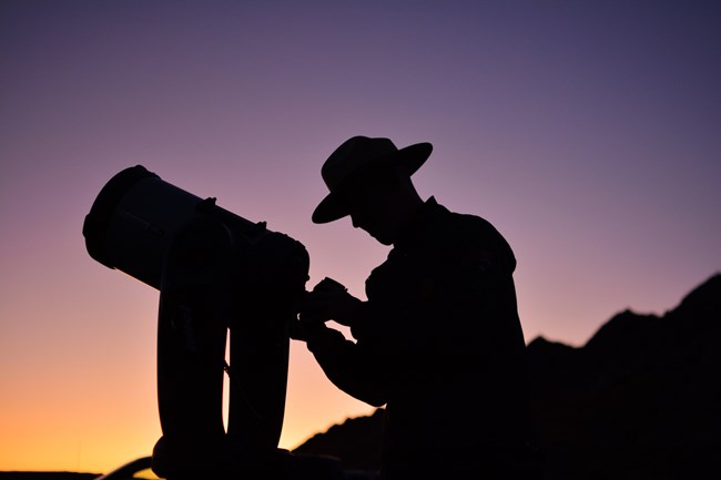 A silhouette of a person setting up a telescope under a pink and yellow sunset. The person is wearing a ranger hat
