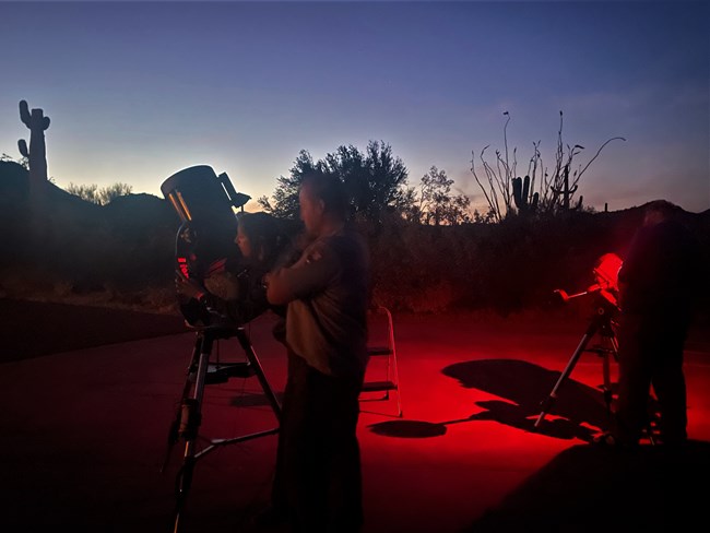 Two rangers stand beside large telescopes under a starry sky