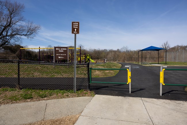 Front view of Bell Acres Park. Entrance features a brown sign that says “Oxon Cove Trail” with a white arrow pointing north. The entrance is gated besides a narrow entrance between a gate. A brown sign that says “Bell Acres Park” is seen behind the gate
