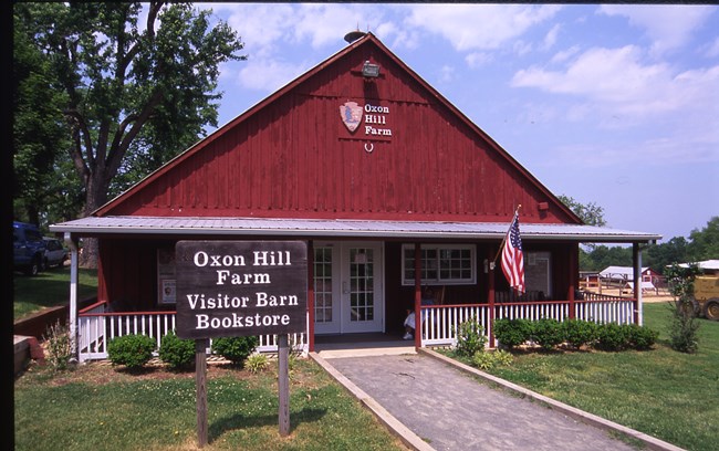 A red barn is positioned in a field. A gravel pathway leads to white double doors. An American flag hangs on the barn.