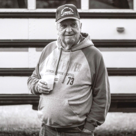 A man stands in front of a school bus with a cup of coffee. His shirt and hat say Jadwin Canoe Rental