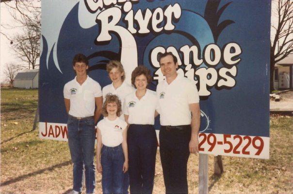 From the 70s or 80s: A family of 5 stands before a small billboard. There is a mother, a father, 2 older sons, and a younger daughter. They all smile brightly at the camera, and they all wear white polo shirts. The sign is a billboard for the Jadwin Canoe