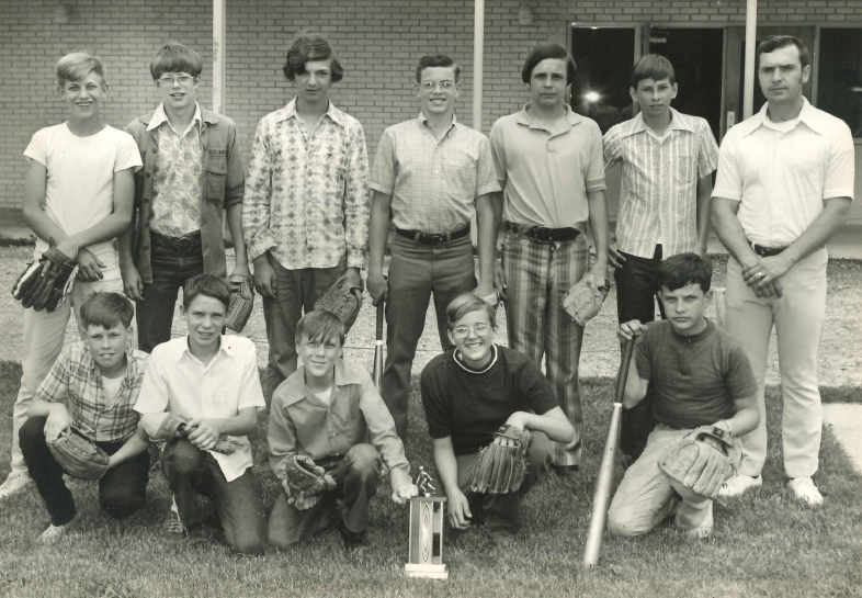 From the 70s or 80s: A group of teenage baseball players pose with their coach. Half kneel in the grass, while the other half stand behind. There is a trophy in front of the team, and various members hold bats and gloves.