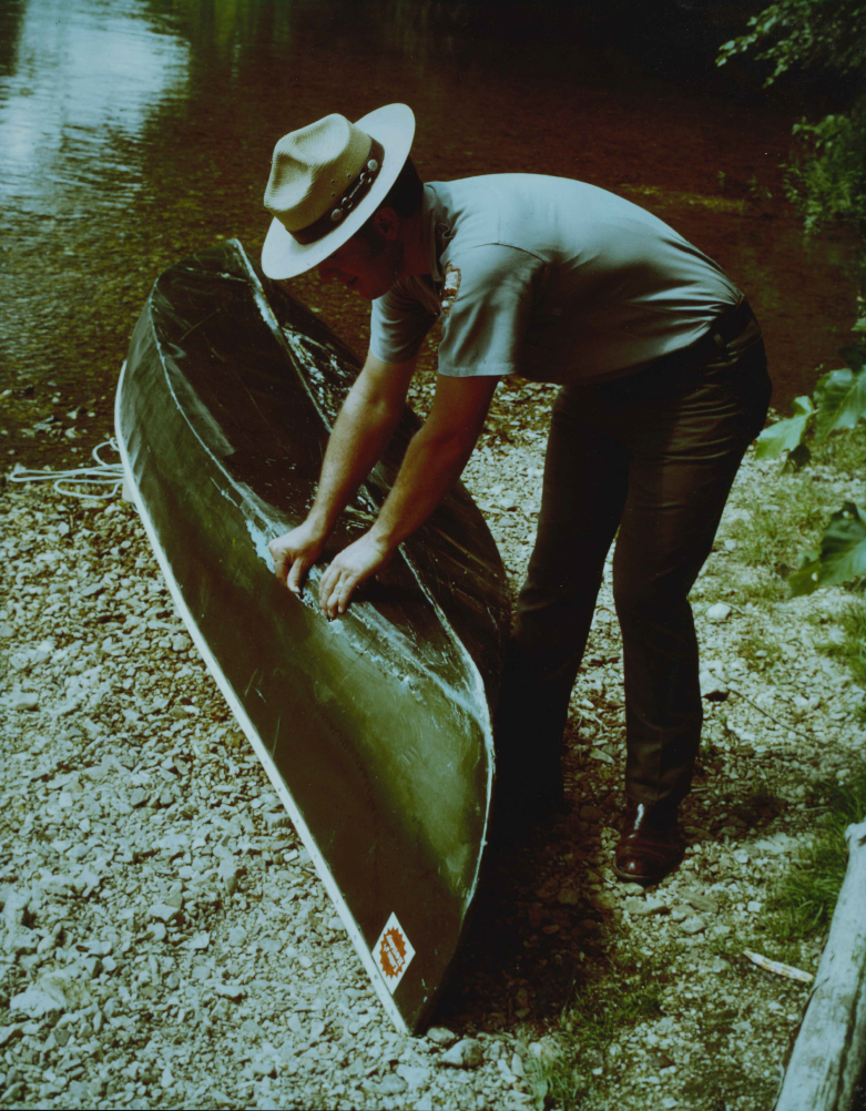 From the 70s or 80s: Darrell Blackwell wears a park ranger uniform as he works on a green canoe. He stands on a gravel bar along the edge of the river. The canoe appears to be torn open by a rock, and Darrell inspects the damage.