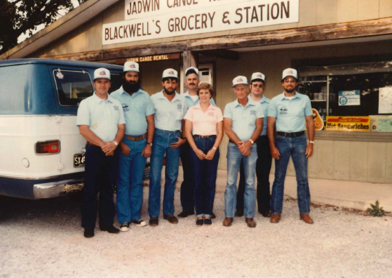 An older image from the 70s or 80s. A group of 7 men and a woman stand before the Jadwin Canoe Rental building. They all wear light blue shirts and blue jeans, apart from the lady, who wears a pink shirt. The men all wear trucker-style hats. A small van c