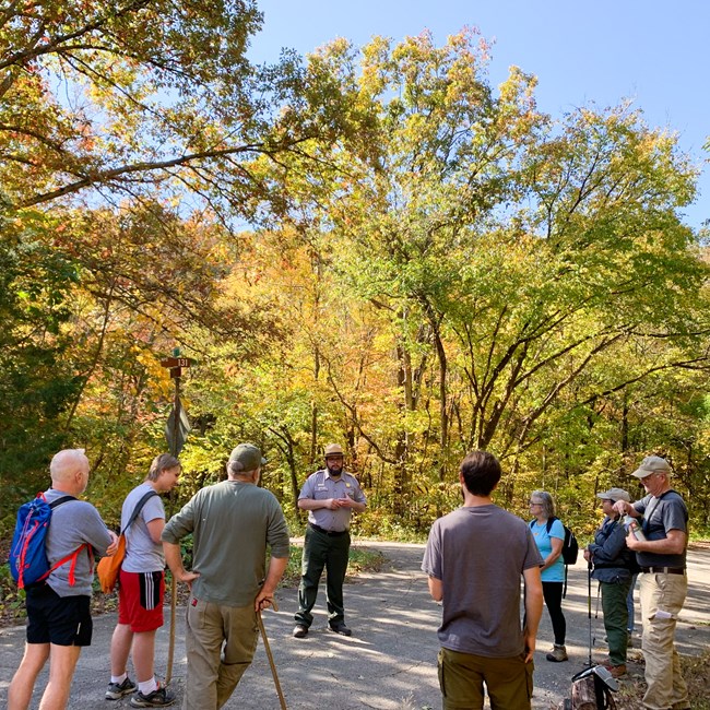A group of hikers stand in front of a ranger. In the background, beautiful fall colors adorn the trees.