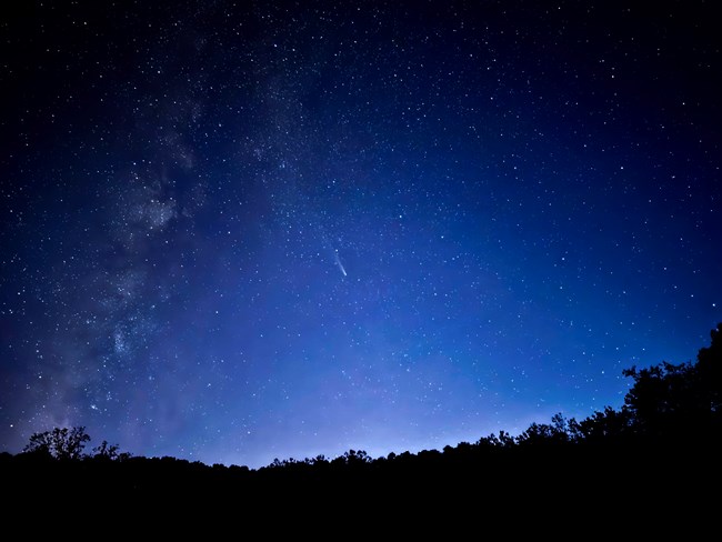 A night sky photo featuring a comet centered on a field of stars. The Milky Way is faintly visible from the lower left to the top center. The bottom of the frame shows a silhouetted horizon with lingering skyglow from sunset, leaving the sky blue-purple..