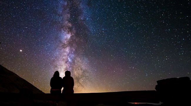 A night sky photograph featuring two people sitting down silhouetted against a dark sky thick with stars. The Milky Way extends straight upwards from the horizon to the top of the frame, where lingering skyglow from sunset is visible.