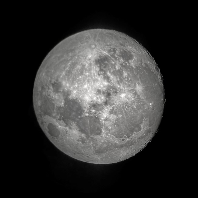 A photograph of Earth's moon taken through a telescope. The moon appears nearly circular with dark grey patches on a lighter grey background.
