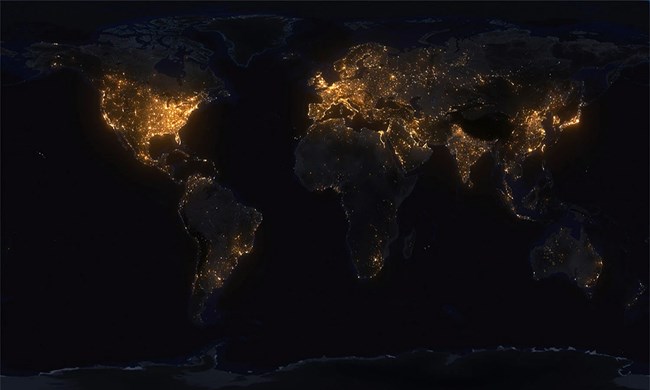 A composite photograph showing the entire world map in darkness with glowing areas of artificial light showing population centers.