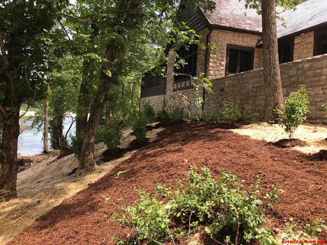 A coursed-cut stone block building with dark brown timber framing and a peaked roof stands on a hillside above a waterway with fresh landscaping dispersed between older trees on the hill below.