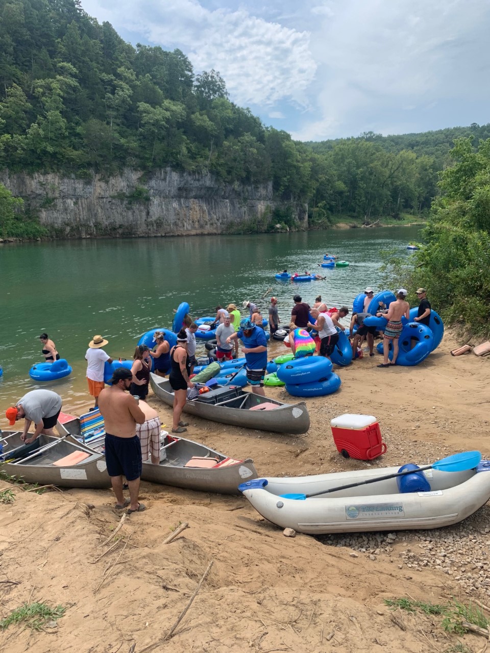 A group of individuals on a river sand bar standing around innertubes, canoes and rafts next to a blue river