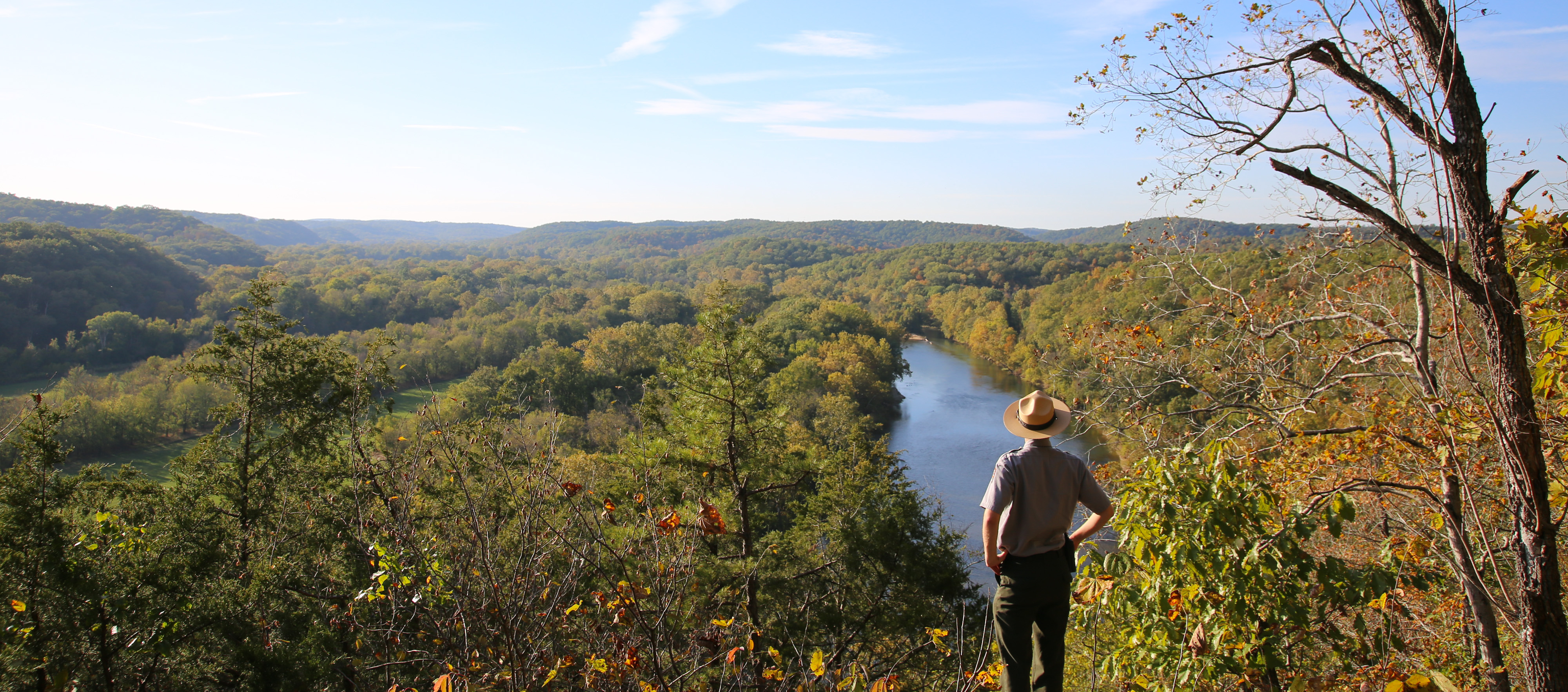 A park ranger stands atop a high cliff, overlooking a river.