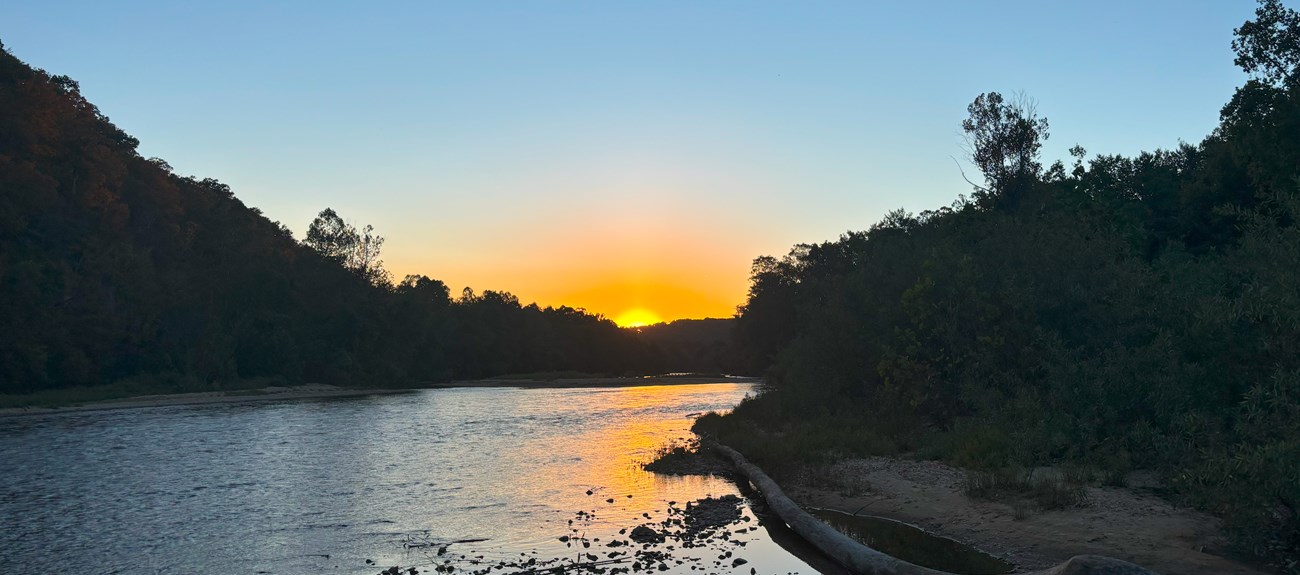 The sun sets along a flowing river.