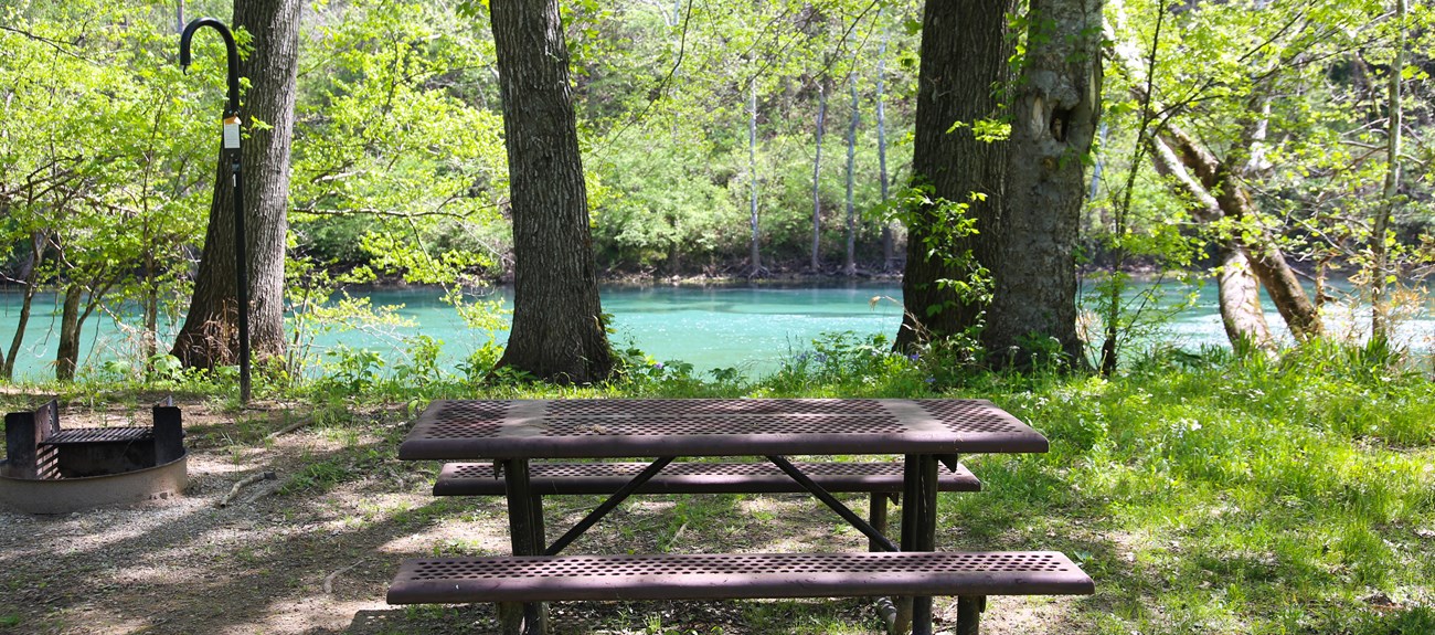 A small campsite sits empty, with a beautiful blue river and green trees in the background.