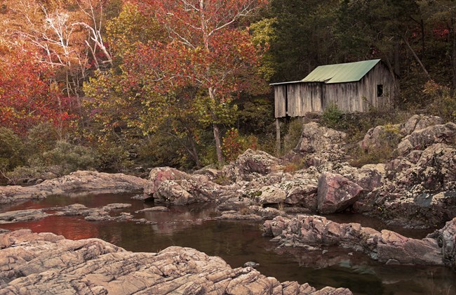 An old metal building with a peaked metal roof sits on the edge of a creek lined with pink, lichen-covered rhyolite boulders with trees in the background beginning to show fall colors.