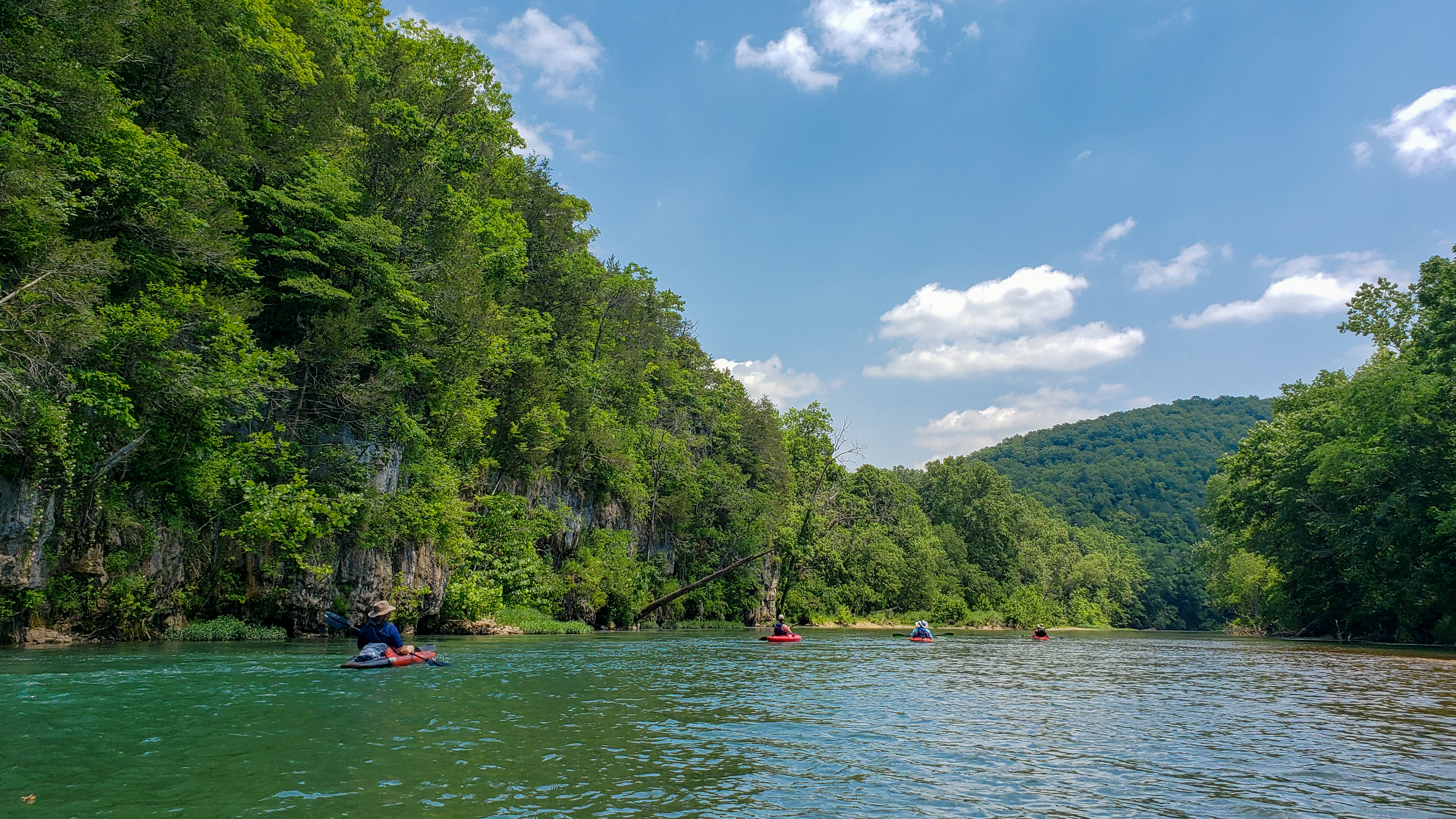 People in red kayaks floating on a calm section of Current River near a forested dolomite bluff.