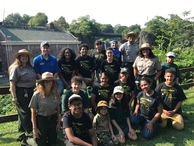 A large group of uniformed park rangers, a blue-shirted Student Conservation Association member, & youths in dark shirts stand before a brick powerplant and 77 ft. waterfall plunging between dark basalt cliffs & an arched black metal bridge