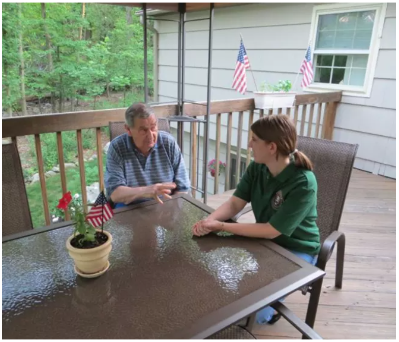 A woman in a green NPS intern shirt sits at a backyard deck table interviewing an older man in a blue & white striped shirt