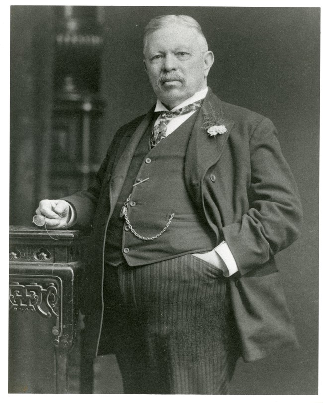 Black & white photo of Catholina Lambert - the slightly rotund, mustachioed silk magnate wears a three piece suit and leans against a table, glasses in hand