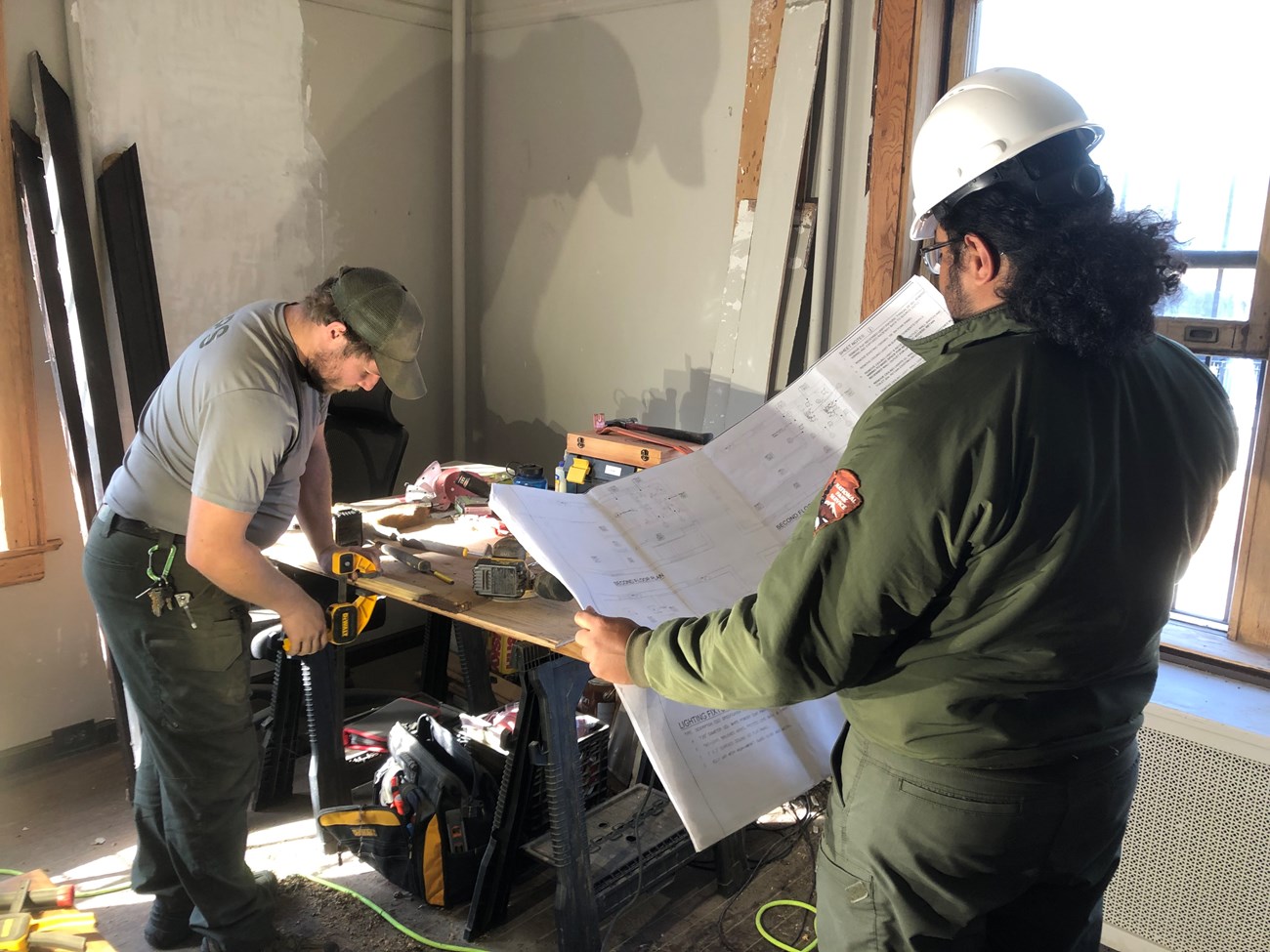 A park ranger in maintenance work clothing works with tools in a room under construction as another ranger in a hard hat consults blueprints