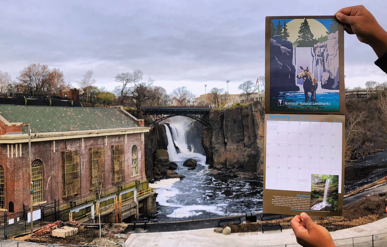 Hands hold a January 2024 National Natural Landmarks calendar before the 77ft. Great Falls, cascading between dark basalt cliffs framed by a black metal arched bridge & a brick hydroelectric power plant