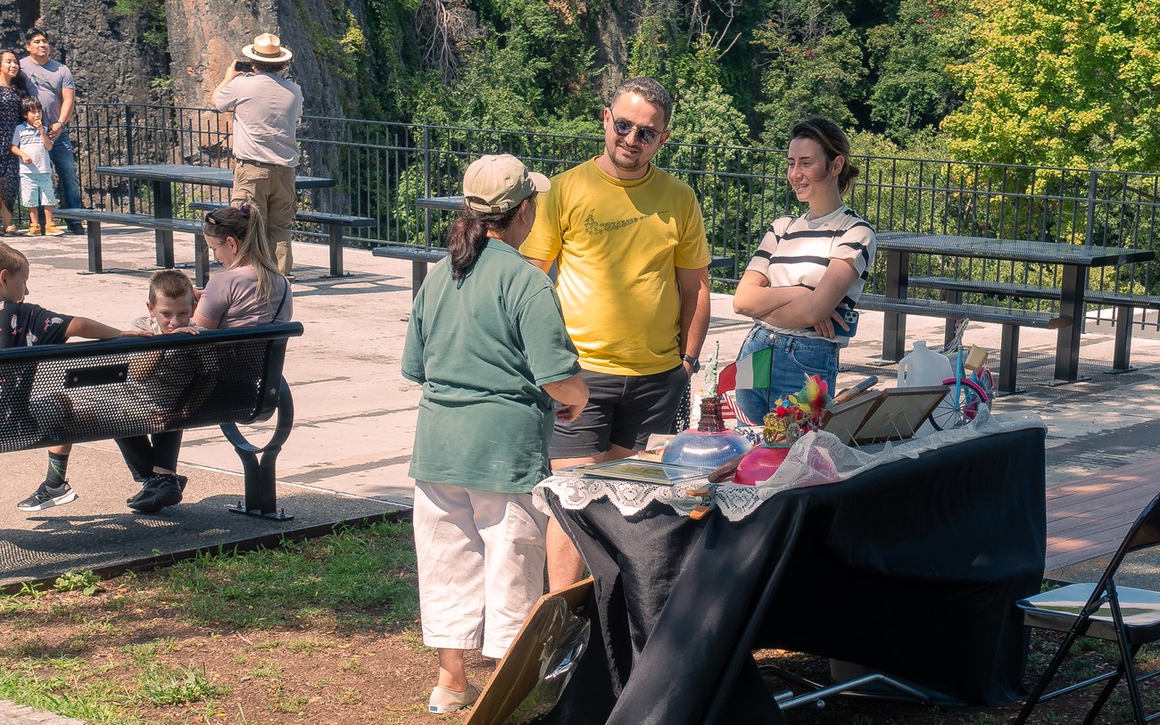 A green-clad volunteer stands before a table of cultural artifacts outdoors, speaking to a pair of visitors