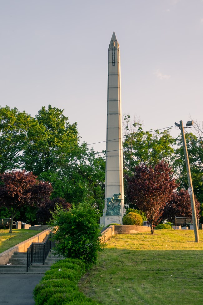 A tall white obelisk with a weathered green copper statue of soldiers at it's base stands in afternoon light at the top of a set of stairs