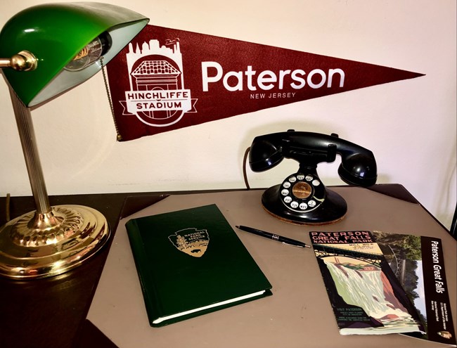 A black rotary telephone sits on a desk beside a green-shaded lamp, a green notebook with a gold NPS arrowhead, a pen, and brochures for Paterson Great Falls. A maroon pennant for Paterson NJ and Hinchliffe Stadium is on the wall.