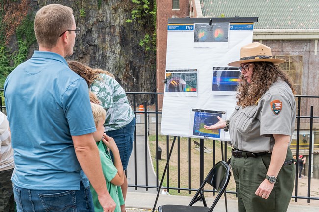 A park ranger gestures towards a science display beside her as a man & a young child listen