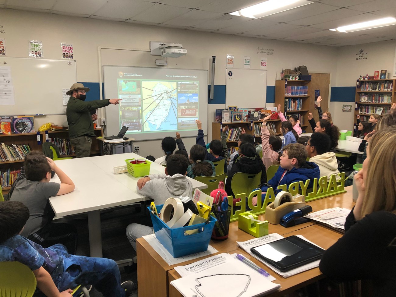Park ranger pointing as 4th-grade students raise hands in a classroom, a powerpoint on a screen