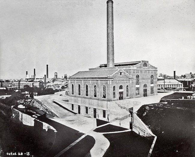 Black & white Oct. 25th 1915 photo of a large brick steam plant - two adjoined buildings w/ tall windows & sloped roofs pierced by a towering chimney. Other mills & chimneys line the horizon