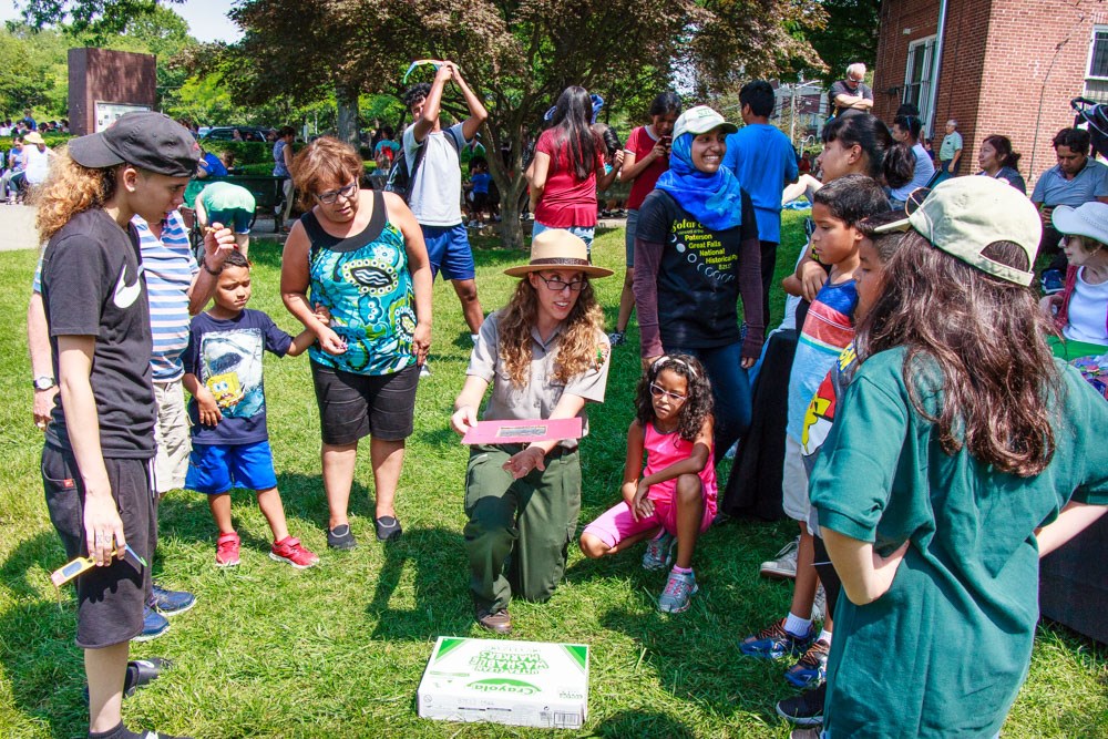 A park ranger demonstrates a flat paper eclipse viewer as children & adults gather around her on grass in front of crowds of people & a shaded area