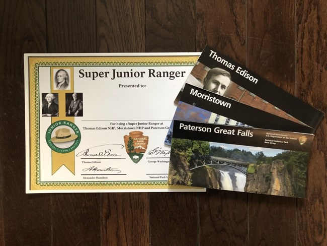 A gold-bordered, glossy Super Jr. Ranger certificate with 3 unigrid brochures from Paterson Great Falls, Morristown, & Edison National Historical Parks sits on a dark wood paneled background