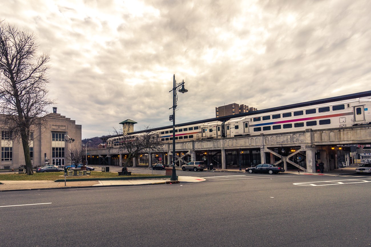 A double-decker NJ Transit commuter train sits in the elevated concrete Paterson train station above the streetscape
