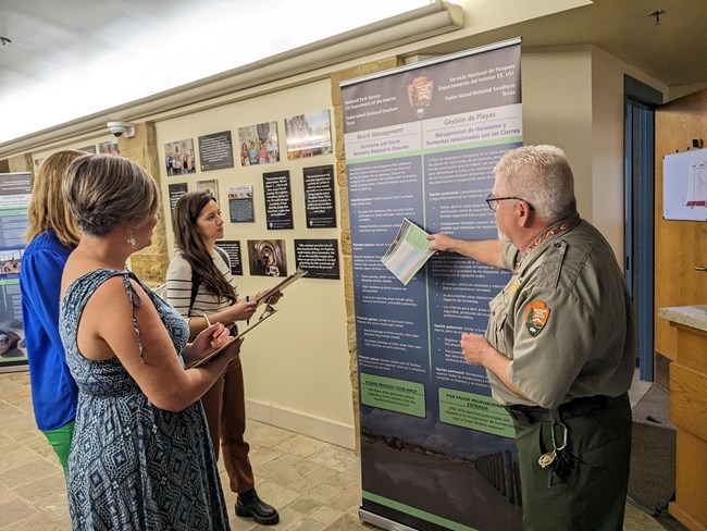 Three people stand around a large poster, a park ranger points twards the poster.