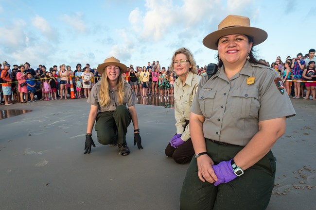 3 rangers kneel on the beach and smile at the camera with a crowd behind them.
