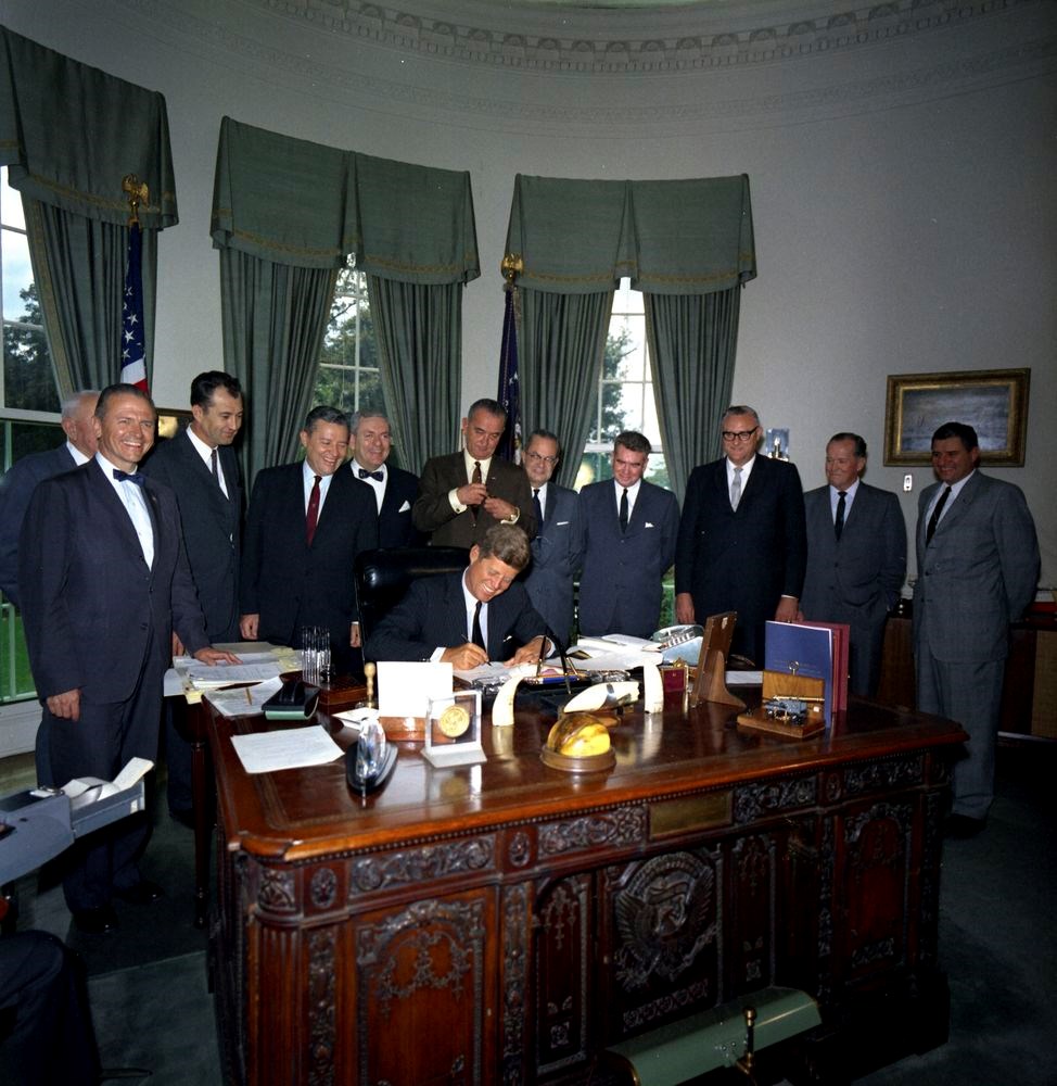 The President Kennedy sitting at a desk signing a bill as others look on.