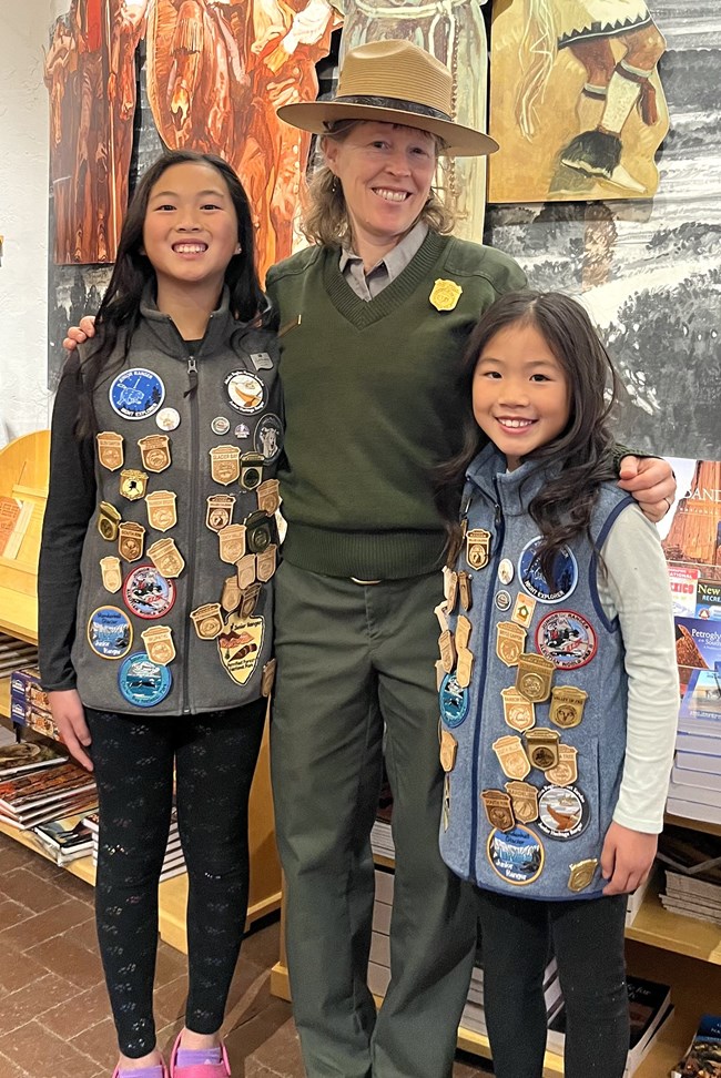 Two children wearing vests covered with junior ranger badges stand next to female park ranger in a bookstore