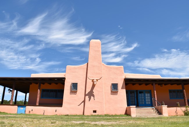 Salmon-colored building with blue trim. Steer-head sculpture attached in the center, shaded porches on each side.