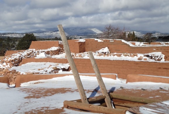 Snow-dusted kiva ladder in front of the adobe walls with clouds and mountains behind.