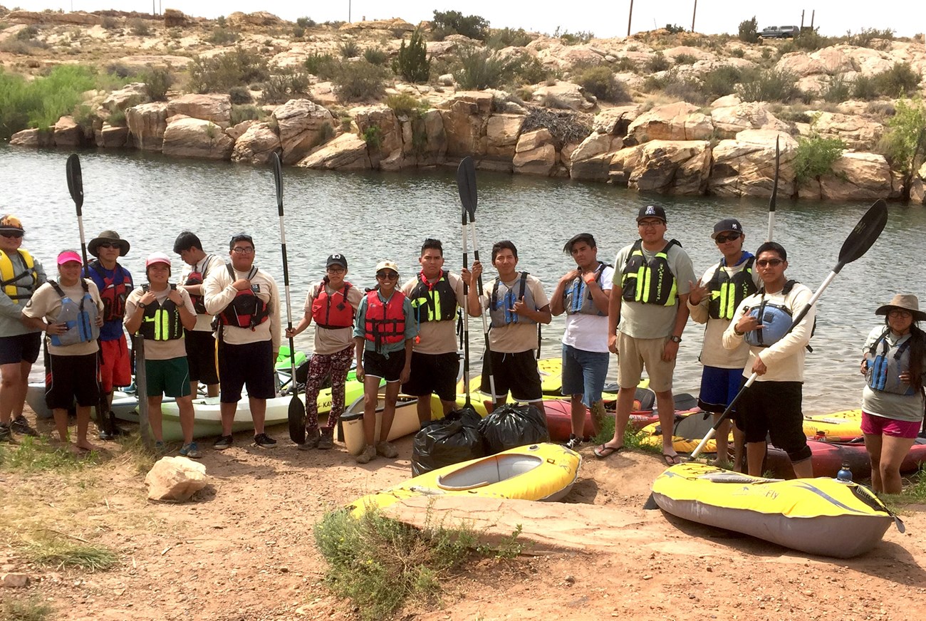 A group of people lined up in front of water and a rock shoreline with paddles and a couple of kayaks.
